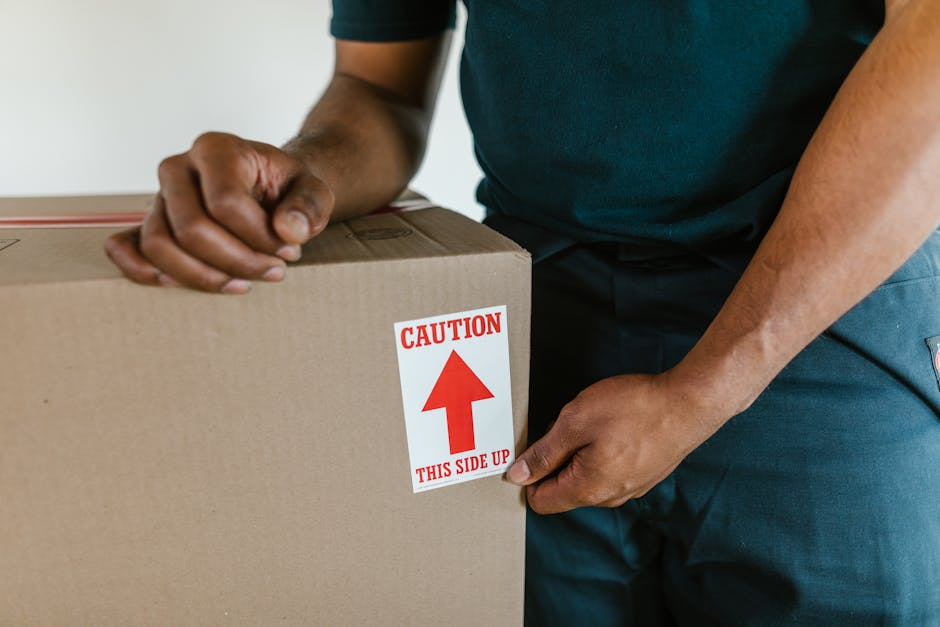 A person wearing a dark green shirt and dark trousers is seen handling a large cardboard moving box indoors. Their hands are positioned on the box, with one hand gripping the side and the other steadying the top. The box features a red and white caution label that reads 'CAUTION THIS SIDE UP' with an upward-pointing arrow, indicating careful handling during the packing and moving process. The surrounding environment includes a clean, well-lit interior space, likely part of a home or rental property, with no other furniture or objects visible. Inside the property, the individual appears to be in the process of packing or preparing items for a home relocation. The image emphasizes the importance of proper packing procedures and careful handling during furniture transport, which are key aspects of professional removals services offered by companies like Man With a Van Harrow.