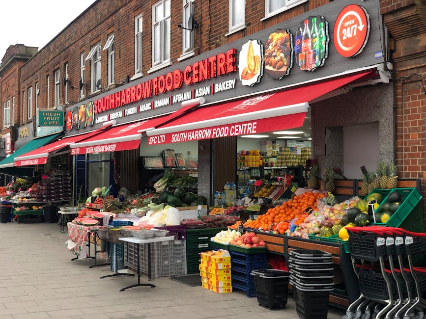 The image shows the exterior of South Harrow Food Centre, a grocery store situated in a brick building with multiple storefronts on a city street. The main signage displays the store's name in large red and white letters, with smaller signs indicating the availability of items such as Turkish, Afghan, Asian, bakery, and halal products. The storefront is partially covered by a red and white awning, which extends over the sidewalk, providing shade. In front of the store, there are several outdoor displays of fresh produce including oranges, watermelons, apples, and various vegetables, arranged in green and black plastic crates, with some also on tables covered with red and white checkered cloths. Additional fruit and vegetables are stacked on the sidewalk, with some in open crates and others in plastic bags. To the left, a small outdoor stall features a variety of flowers in pots, along with freshly picked fruit. The scene is captured during daylight under natural lighting, portraying a typical urban environment where [COMPANY_NAME] might assist with home relocation or furniture transport involving packing and moving processes, especially when planning for house removals or moving logistics.