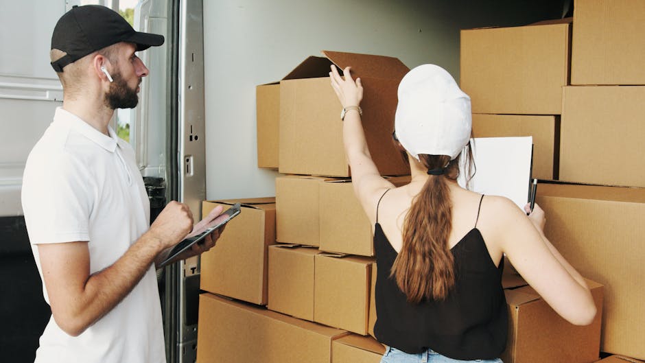 A person wearing a dark green shirt and dark trousers is seen handling a large cardboard moving box indoors. Their hands are positioned on the box, with one hand gripping the side and the other steadying the top. The box features a red and white caution label that reads 'CAUTION THIS SIDE UP' with an upward-pointing arrow, indicating careful handling during the packing and moving process. The surrounding environment includes a clean, well-lit interior space, likely part of a home or rental property, with no other furniture or objects visible. Inside the property, the individual appears to be in the process of packing or preparing items for a home relocation. The image emphasizes the importance of proper packing procedures and careful handling during furniture transport, which are key aspects of professional removals services offered by companies like Man With a Van Harrow.