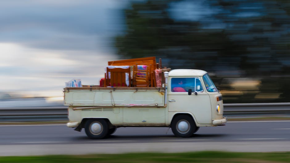 A vintage cream-colored three-wheeled van with a flatbed, used for home relocation or moving services, is shown in motion on a road with a blurred background of trees and sky, carrying various household furniture and packed boxes. The load includes a wooden dining chair, a small wooden table, and several cardboard boxes wrapped in plastic, secured on the flatbed with straps. The van's interior cabin is partially visible, with the driver’s side mirror and window. The image captures the vehicle during the loading process or transport stage of a house removal, highlighting the challenge of bulky item access on Harrow estates and the need for professional removals solutions, as offered by Man With a Van Harrow.