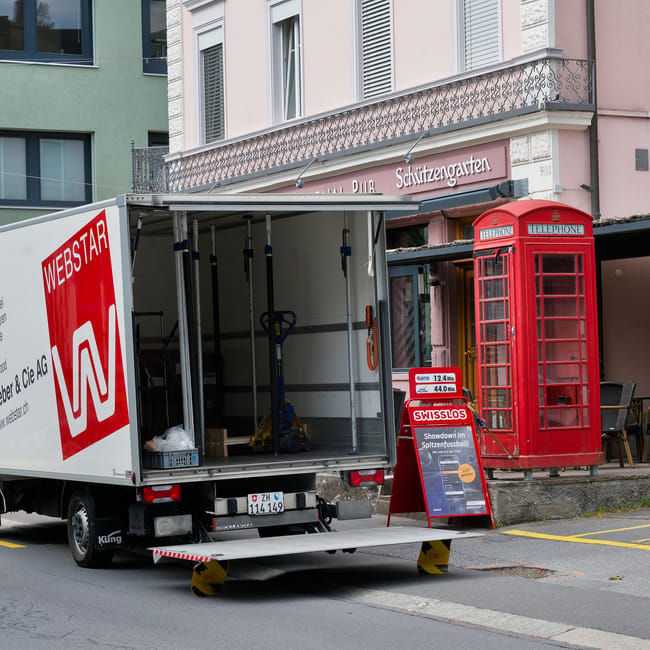 A white moving van with the side door open, parked on a city street in front of a building with a pink and white facade and a decorative balcony. Inside the van, there are visible tools and equipment used for home relocation, including a hand truck, orange straps, and packing supplies. The rear of the van is equipped with a loading ramp extended onto the pavement, assisting the loading process of large furniture or appliances. Nearby, there's a traditional red telephone booth and a black outdoor seating area, with the van positioned directly in front of a café or pub, indicated by signage for 'Schutzengarten.' The scene captures the logistical stage of furniture transport and packing during a house move, supported by the services of Man With a Van Harrow, aimed at addressing bulky item access problems on Harrow estates and providing efficient removals solutions.