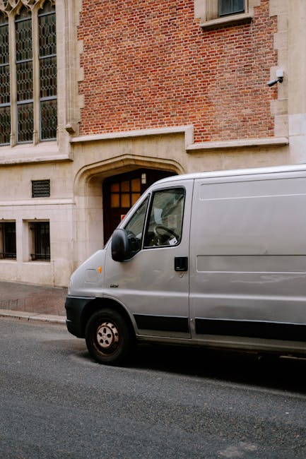 A silver van from Man With a Van Harrow parked on a street in front of a historic red brick building with large stained-glass windows and a stone arch doorway. The van is positioned close to the building's entrance, partially on the pavement, during daylight with natural lighting. The vehicle is used for home relocation or furniture transport, typical of removals services, and is situated near a curb on an urban street. The background includes the building’s stone and brick façade, with some small windows and a security camera mounted on the wall. This scene illustrates the loading process during a house move, with the van ready for carrying packed boxes and furniture into or out of the property, supporting the logistics of a professional removals company like Man With a Van Harrow, providing essential moving and packing services.
