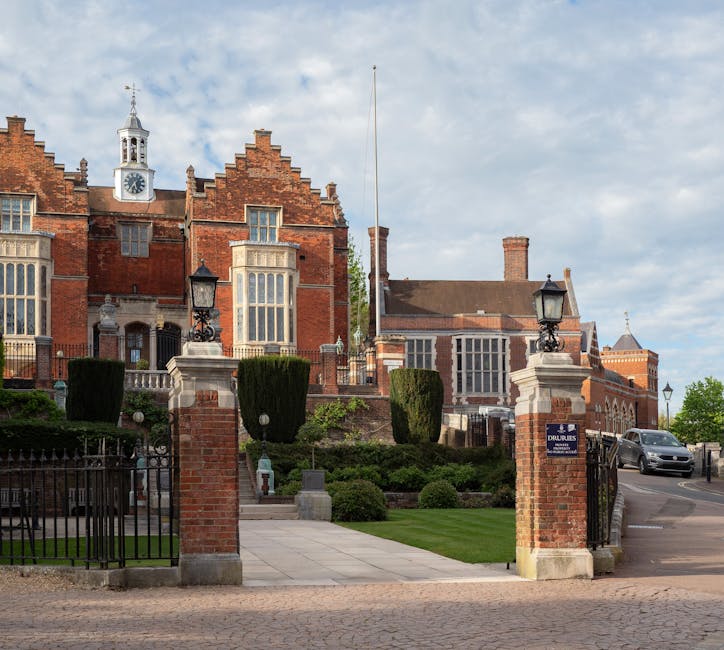 A view of a large, historic brick building with a clock tower and decorative gables, surrounded by a well-maintained garden with neatly trimmed bushes and tall, dark green hedges. The entrance is marked by a set of brick pillars topped with black wrought iron lamp posts, and a black iron gate partially open to reveal a paved pathway leading toward the building. To the right, a silver van is parked on the street near the entrance, highlighting the context of furniture transport and home relocation logistics. The scene is illuminated by natural daylight with a partly cloudy sky overhead, suggesting daytime loading or unloading activities as part of a house removal process facilitated by Man With a Van Harrow.