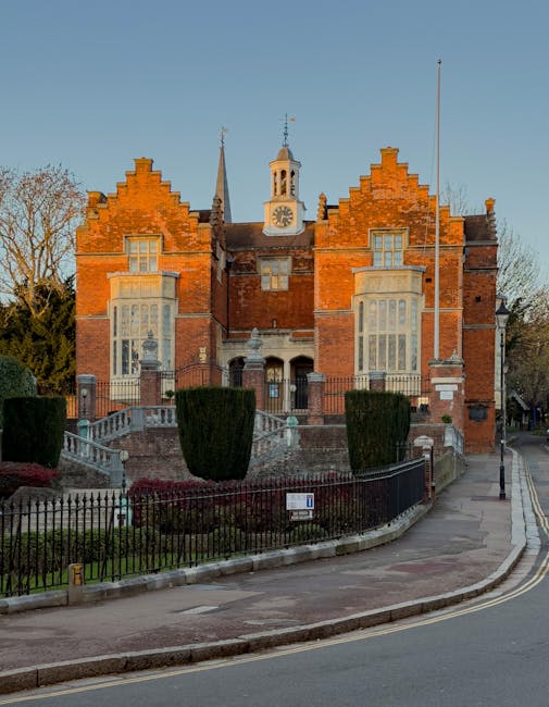 A silver van from Man With a Van Harrow parked on a street in front of a historic red brick building with large stained-glass windows and a stone arch doorway. The van is positioned close to the building's entrance, partially on the pavement, during daylight with natural lighting. The vehicle is used for home relocation or furniture transport, typical of removals services, and is situated near a curb on an urban street. The background includes the building’s stone and brick façade, with some small windows and a security camera mounted on the wall. This scene illustrates the loading process during a house move, with the van ready for carrying packed boxes and furniture into or out of the property, supporting the logistics of a professional removals company like Man With a Van Harrow, providing essential moving and packing services.