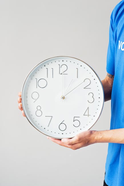 A person dressed in a blue uniform holding a large round wall clock with a white face, black numerals, and minimalistic black hands. The individual is standing indoors against a plain gray background, with their left hand supporting the bottom of the clock and their right hand gripping the side. The clock shows the time as approximately 12:10. The scene is related to home relocation and furniture transport, with the clock representing time-sensitive moving schedules typical of last-minute Harrow removals, as offered by Man With a Van Harrow. The setting emphasizes careful handling of belongings during packing and loading processes, highlighting the importance of efficient and professional removals services.