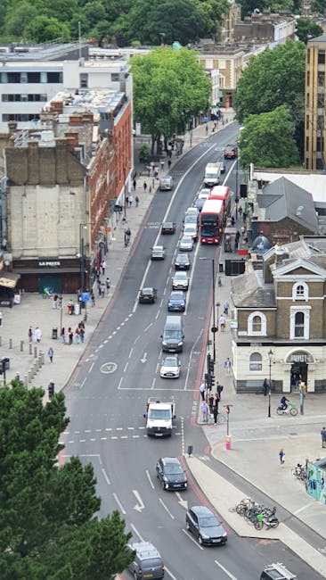 An aerial view of a busy urban street during daytime, showing a line of vehicles including cars and buses moving along a curved road bordered by commercial and residential buildings. On the left, pedestrians walk on the sidewalk, some crossing at the designated crosswalks. The scene features a mixture of modern and older architecture, with trees providing greenery along the street. In the foreground, a delivery van, associated with [COMPANY_NAME], is parked near a building, while residents and movers may be seen navigating the area. The overall environment suggests active city life with vehicle and foot traffic typical of a central area in Harrow, supporting the context of home or furniture transport, packing, and moving logistics in relation to [PAGE_TITLE]. The lighting is natural and bright, highlighting the details of the street scene, which is relevant for visualizing urban moving and relocation services provided by [DOMAIN].