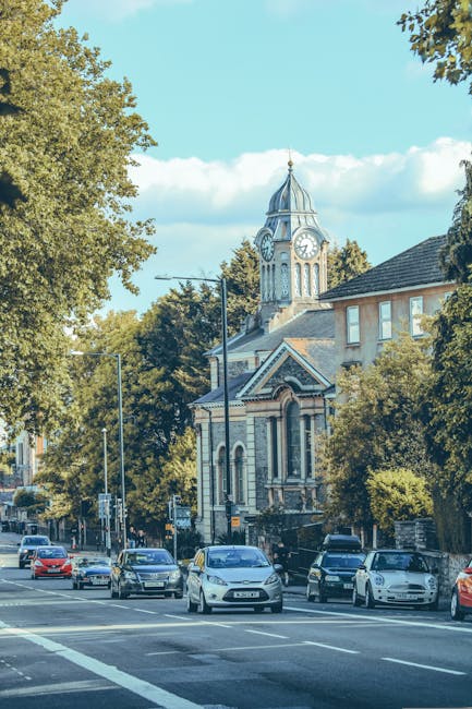 A street scene in Harrow with several parked and moving cars on a paved road, bordered by tall leafy trees on each side. In the background, a historic church building with a clock tower and ornate architecture is visible, partially obscured by the trees. The scene is captured in daylight with a clear blue sky and some clouds. Although no furniture, boxes, or packaging materials are shown in this image, Man With a Van Harrow offers professional house removals and furniture transport services, often involving loading and unloading processes in similar urban environments, ensuring efficient home relocation and moving logistics.