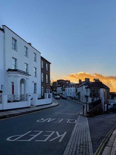 A quiet residential street scene at sunset with a gentle incline, featuring white and dark brick terraced houses on both sides. The street is paved with asphalt, marked with a large white 'KEEP CLEAR' warning painted on the road surface, and has a tactile paving strip along the sidewalk edge for accessibility. The houses have small front gardens and balconies, with some displaying decorative railings. In the foreground, a section of a man with a van from Man With a Van Harrow is visible, indicating a home relocation or furniture transport process. The sky is clear with a few orange and yellow clouds near the horizon, casting warm light over the street, suggesting a typical moving or packing day during late afternoon or early evening.