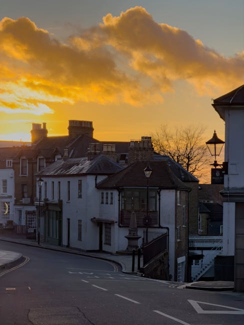 A street scene during sunset showing a row of residential buildings with varying architectural styles, including brick and white painted facades, some with chimneys and bay windows. The pavement is vi