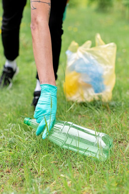A close-up view of a person wearing a blue disposable glove picking up a large, empty plastic bottle from a grassy outdoor area during a cleanup activity. In the background, there is a yellow and tran