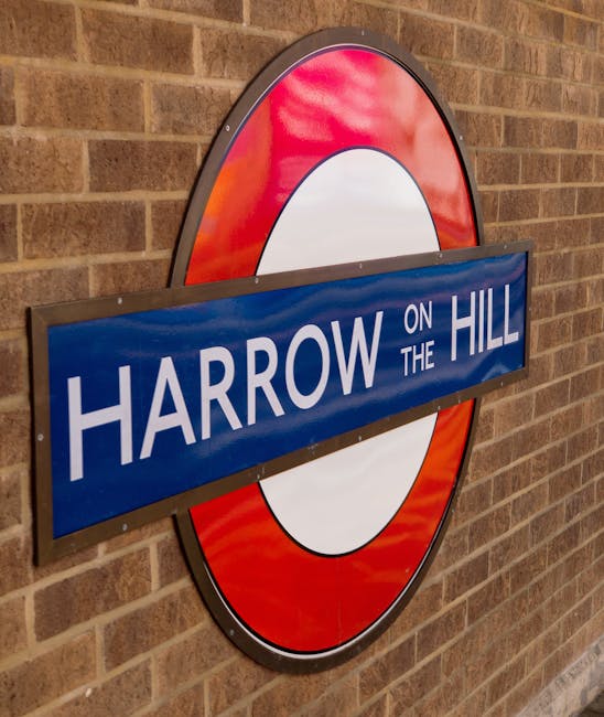 A close-up image of a round London Underground-style sign mounted on a brick wall, featuring a red circular border and a white center. A blue rectangular nameplate with white uppercase letters spelling 'HARROW ON THE HILL' is horizontally affixed across the sign. The sign is outdoors, adjacent to a building or station platform, and appears to be associated with local transport or signage indicating a station or area within Harrow. In the context of house removals and moving services, this type of signage is relevant for property location identification, which Man With a Van Harrow might coordinate with during home relocation or furniture transport to or from the Harrow area.