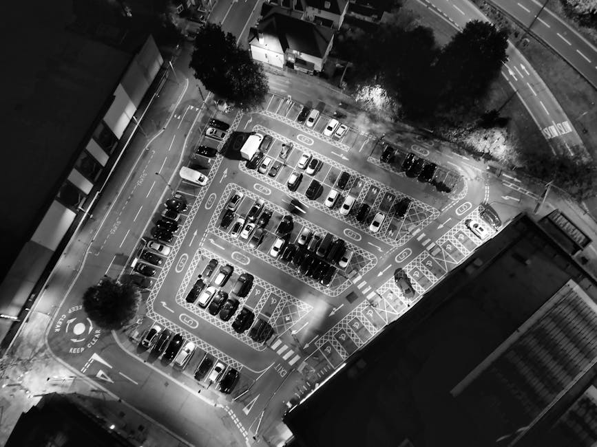 Black and white aerial photograph of a multi-storey car park at night, showing parked cars in designated spaces, some lights illuminating the area, and trees lining the perimeter. The parking lot has clearly marked parking bays, directional arrows, and signage, with a surrounding urban environment including adjacent buildings and roads. The image captures the organized layout of vehicle storage, which is relevant for home relocation and furniture transport services provided by Man With a Van Harrow, illustrating the logistical aspects of loading and unloading during house removals and moving processes.